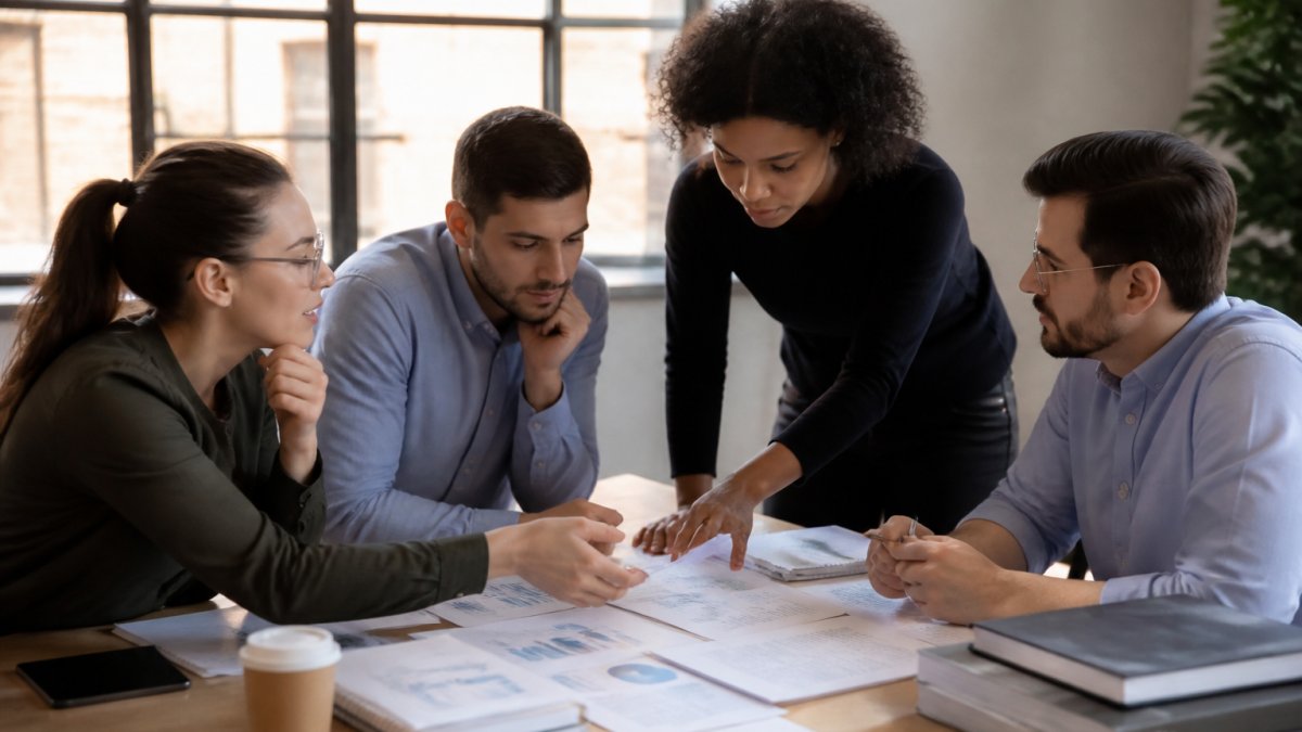 Professional research team reviewing brand analytics on multiple monitors in collaborative workspace, discussing data reconciliation and cross-checking findings with natural office lighting